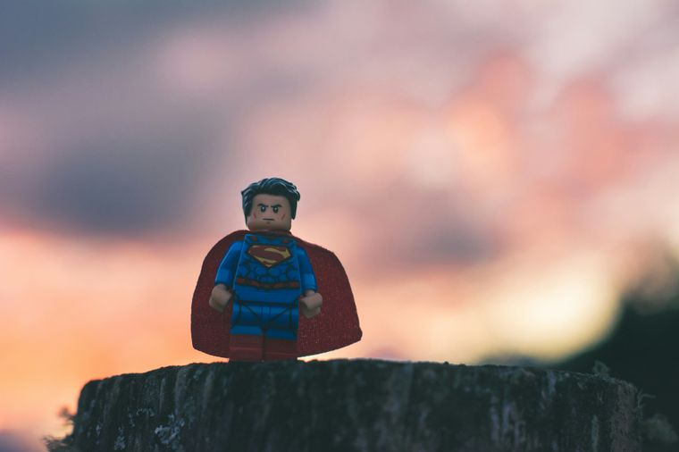 A Lego superman figure with their cape flying in a wind in front of a dramatic orange/pink sky